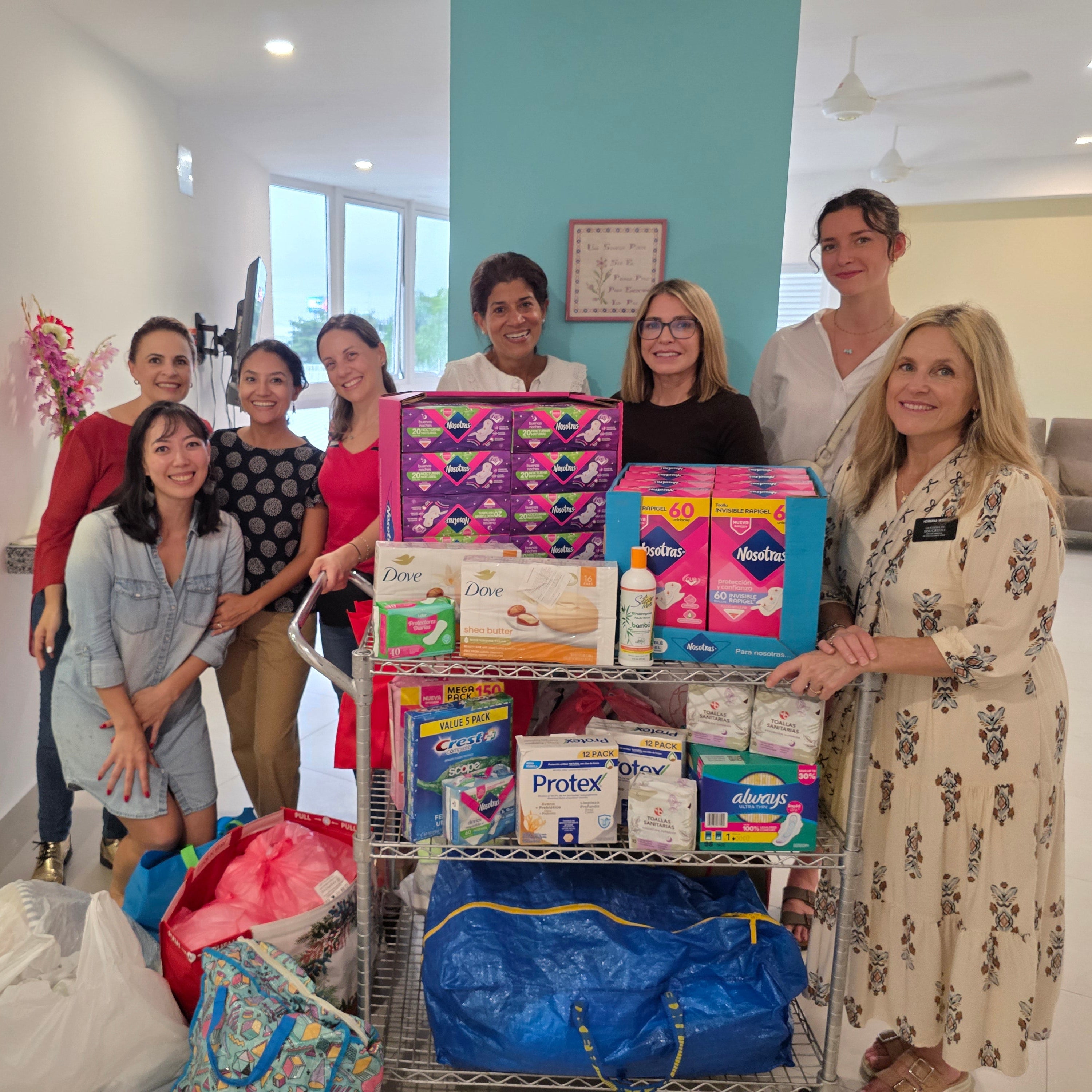A group of eight women standing behind a wire utility cart loaded with packaged household goods, diapers, laundry detergent, and bags in a brightly lit indoor hallway.