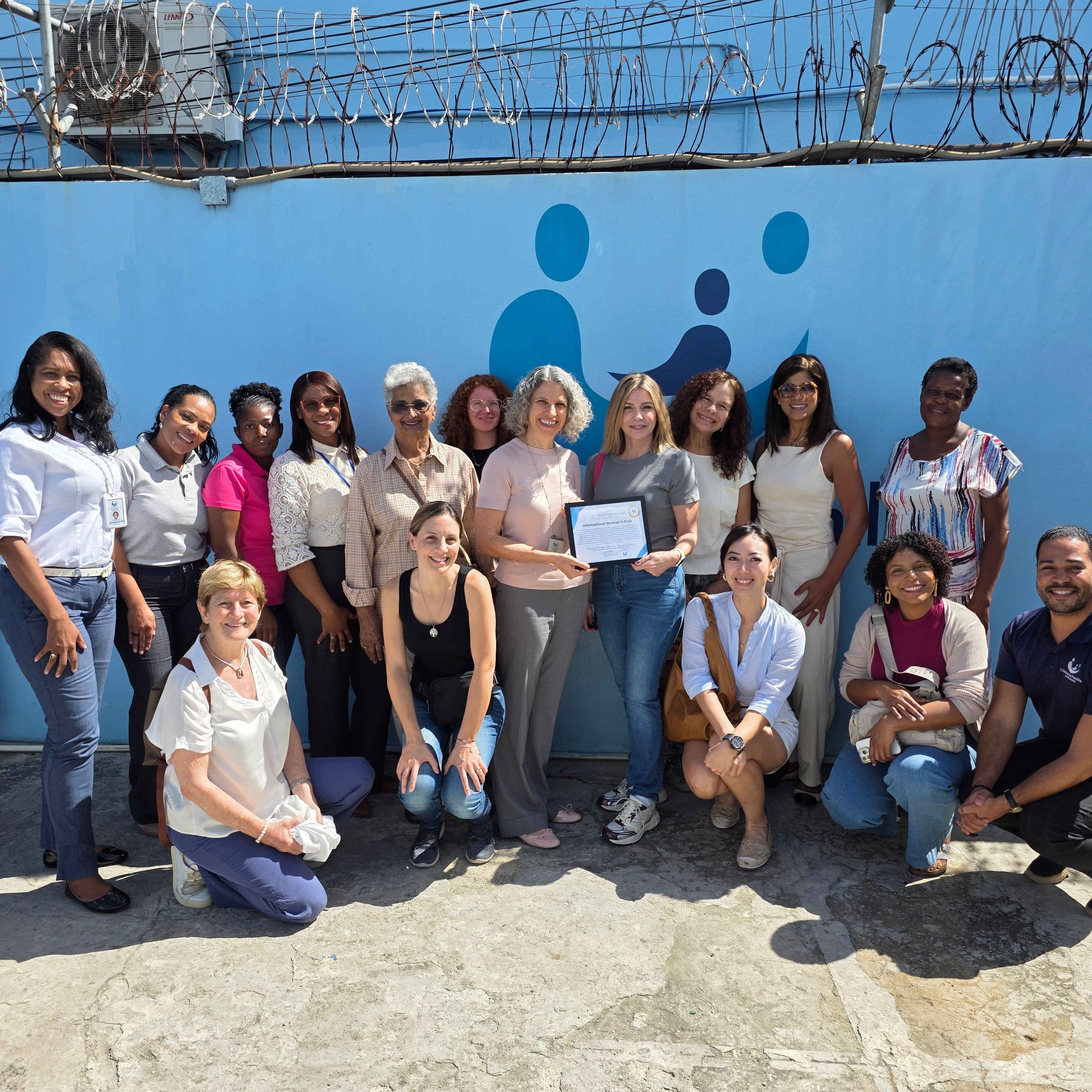 A group of fifteen people posing for a group photo in front of a light blue wall with barbed wire on top. The group is smiling and holding a small certificate.